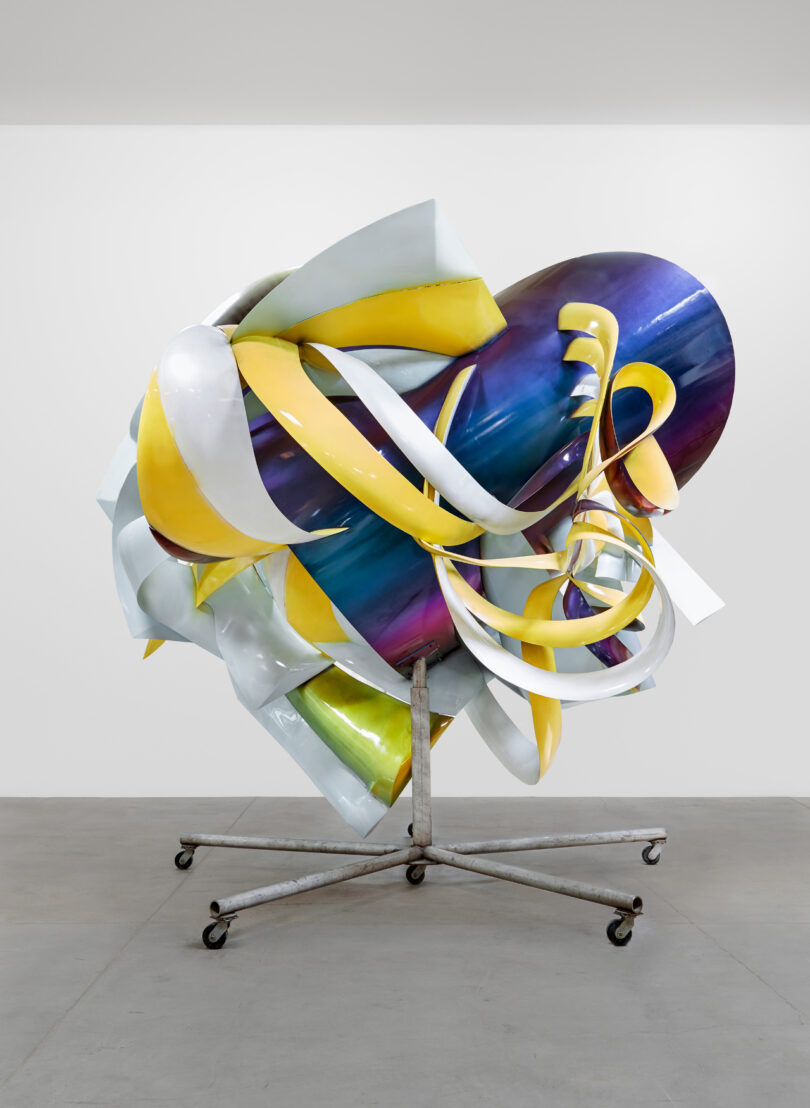 Colorful, abstract metal sculpture on a wheeled stand, displayed in a white gallery space.
