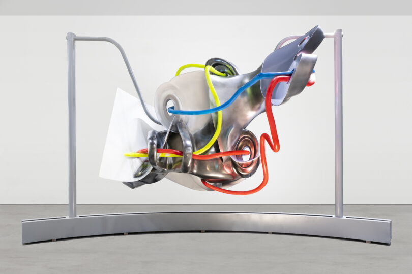 Abstract metal sculpture with colorful tubing elements, displayed on a minimalist frame in a gallery setting.