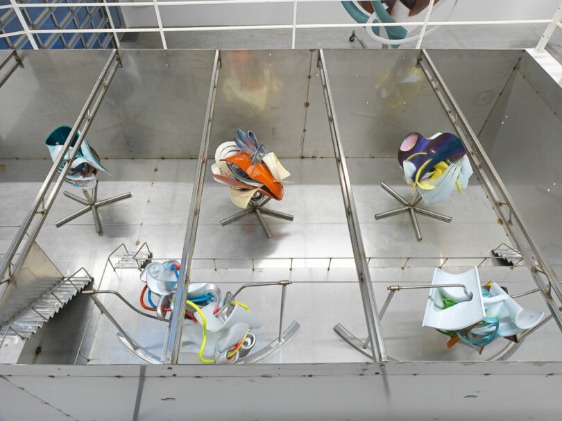 Overhead view of colorful modern chairs scattered on glass flooring with metal supports and stairs visible.