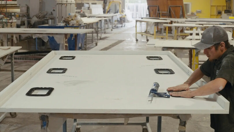 A man working on a white stone countertop installing six wireless charging systems onto the underside of the surface in a factory setting.