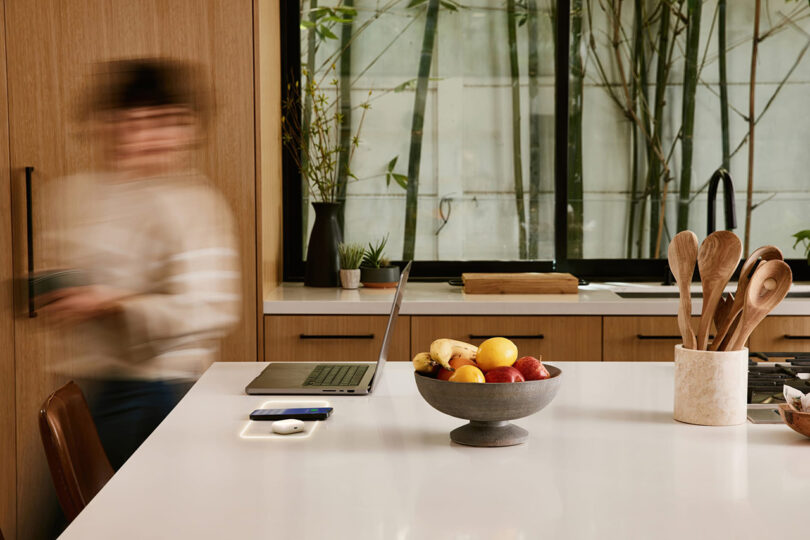 A woman in blurred motion in a kitchen with a laptop on a wireless charging pad.