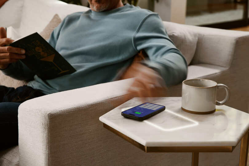 A man sitting on a couch, enjoying a book while his phone is on wireless charging.