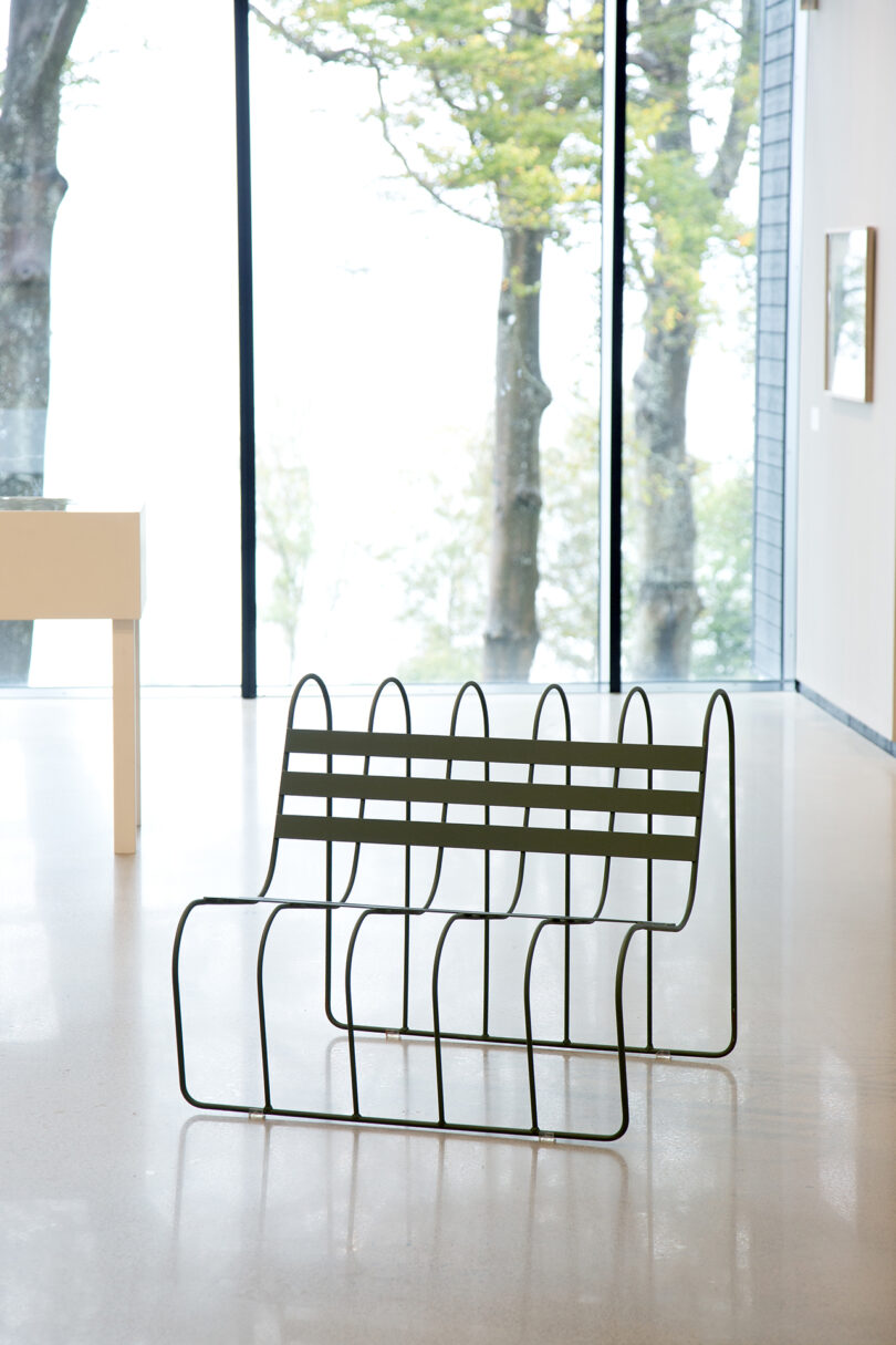 A minimalist metal bench situated in a bright room with a large window offering a view of trees.