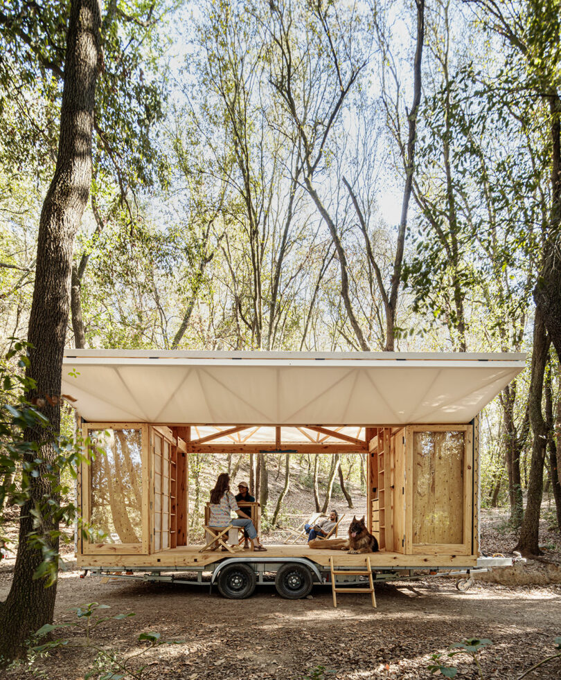 A portable wooden structure built on a trailer parked in a forest setting with individuals relaxing inside.