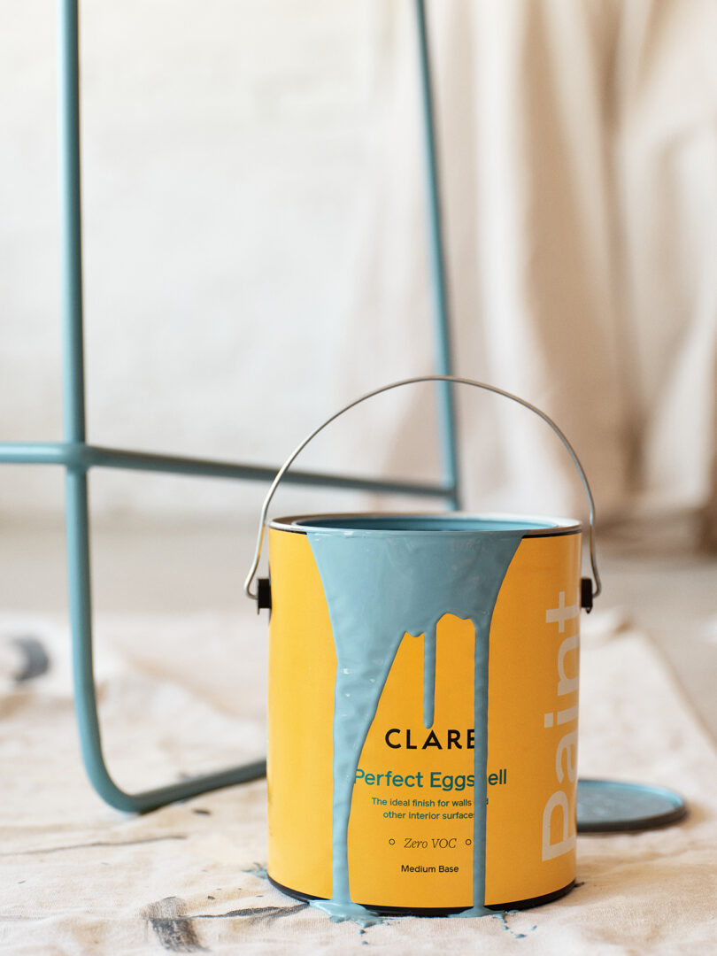 A paint can with blue paint dripping down the side stands on a drop cloth-covered floor.