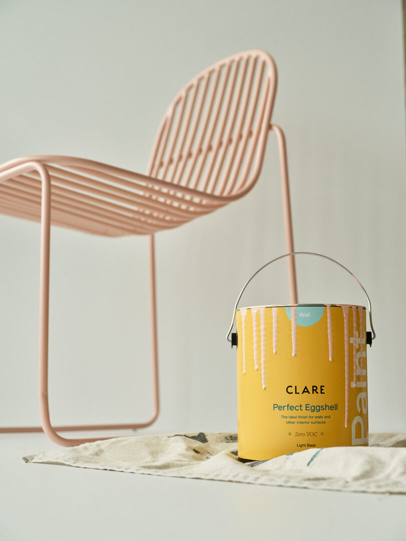 A pastel pink chair next to a yellow can of paint with paint drips on a drop cloth.