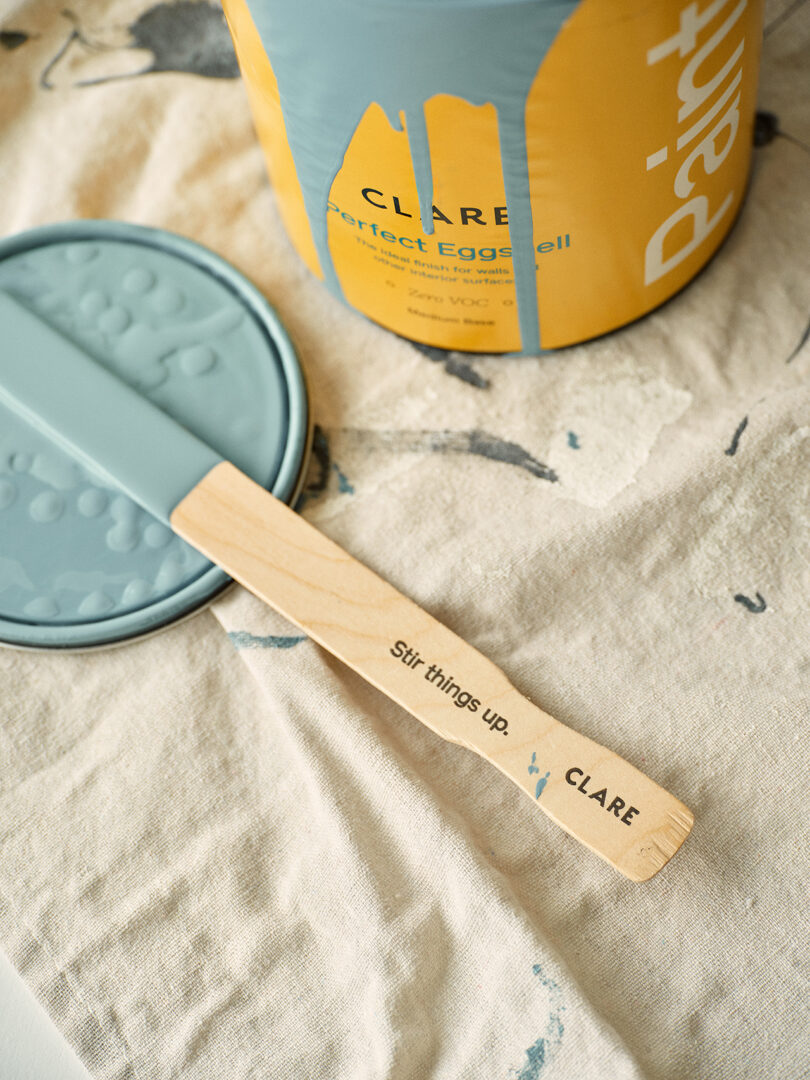 A paint stirrer with the phrase "stir things up" rests on a canvas, next to a paint can with a lid.