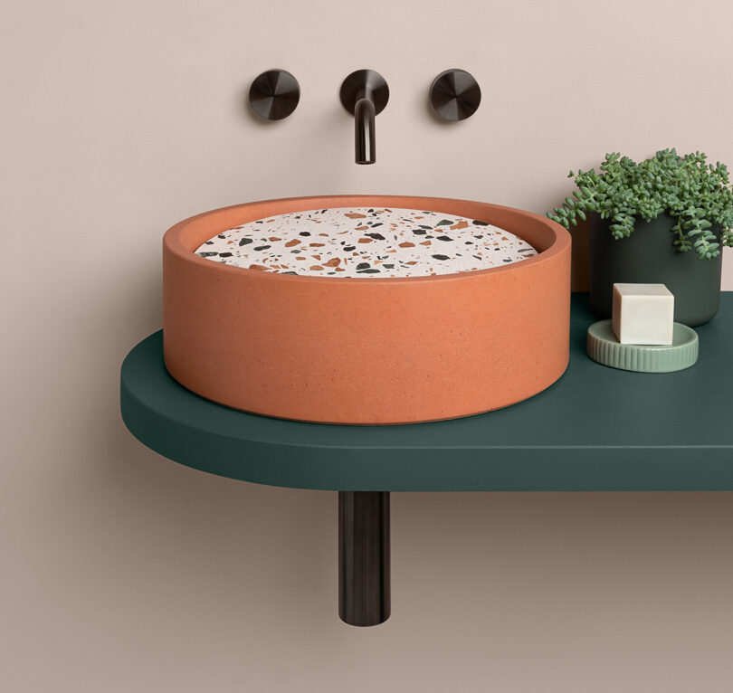 A clay colored concrete bathroom sink with terrazzo insert with a potted plant on the side and small soap dish. Sink faucet and dials are black and wall mounted on a light pink wall.