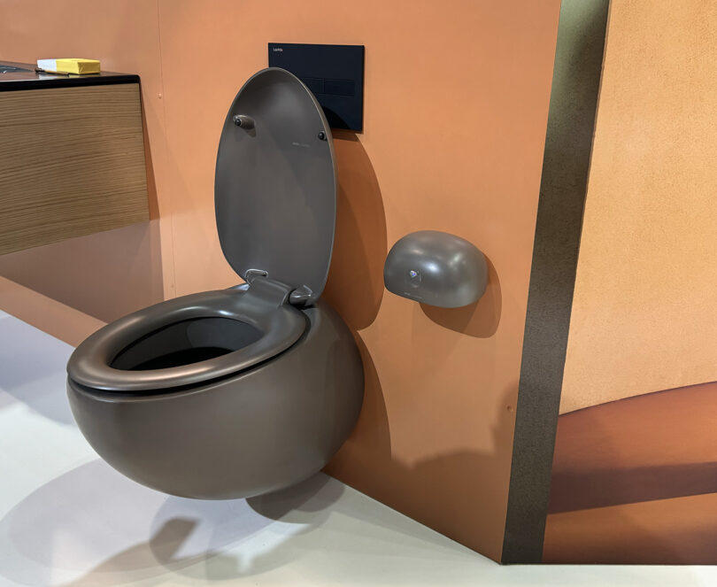 A latte-colored wall mounted toilet attached to a rust hued wall at KBIS.