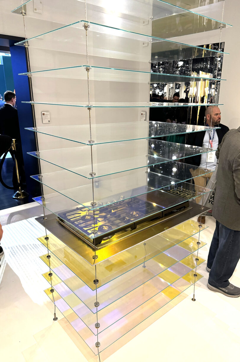 A tiered clear shelf display with a gas cooktop installed on the 5th level of glass. Two men are standing to the right side alongside the display.
