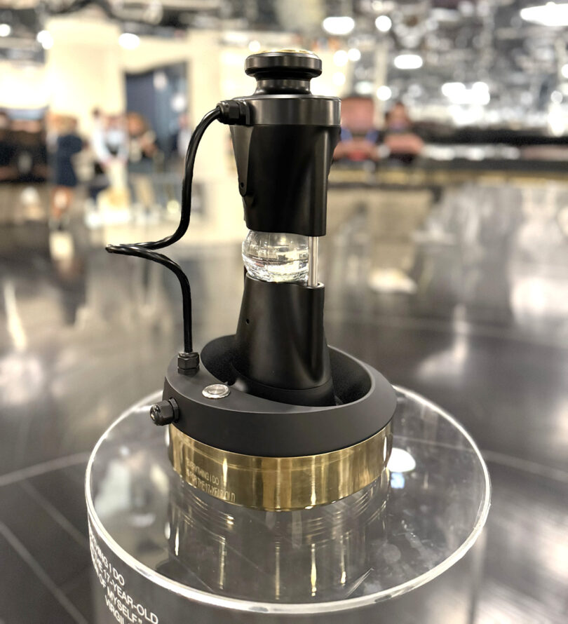 A black and gold sphere ice making device set on a clear surface pedestal