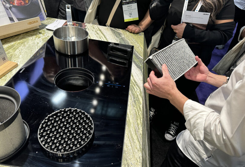 A person holding a BORA kitchen air extraction filter with small crowd of KBIS attendees standing around an induction cooktop with pot boiling nearby.