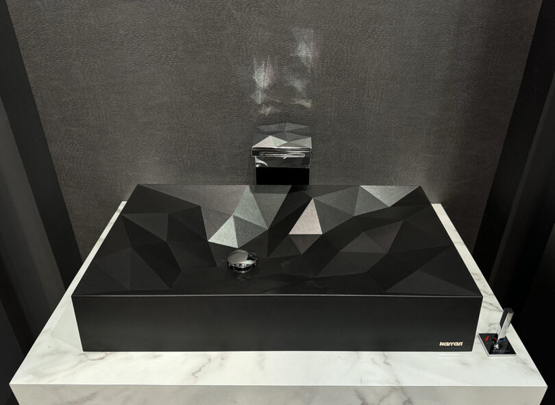 A black faceted rectangular sink with a silver faucet featured at KBIS on a marble pedestal
