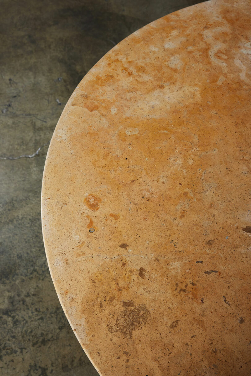 View of a round stone tabletop with a stained and textured surface against a concrete floor beneath.
