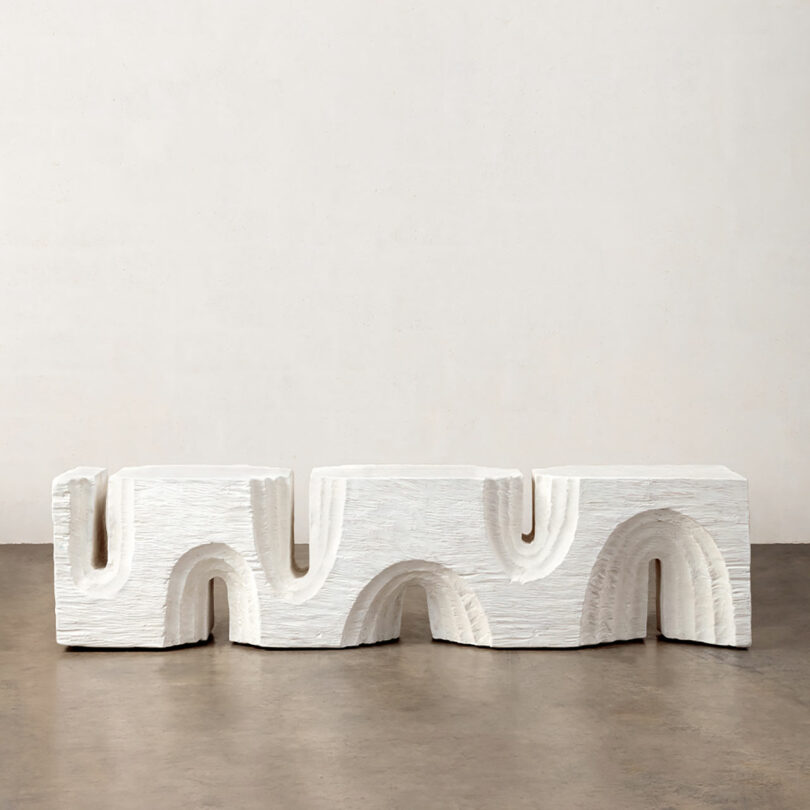A white, wavy modular bench structure on a gallery floor with a plain wall in the background.