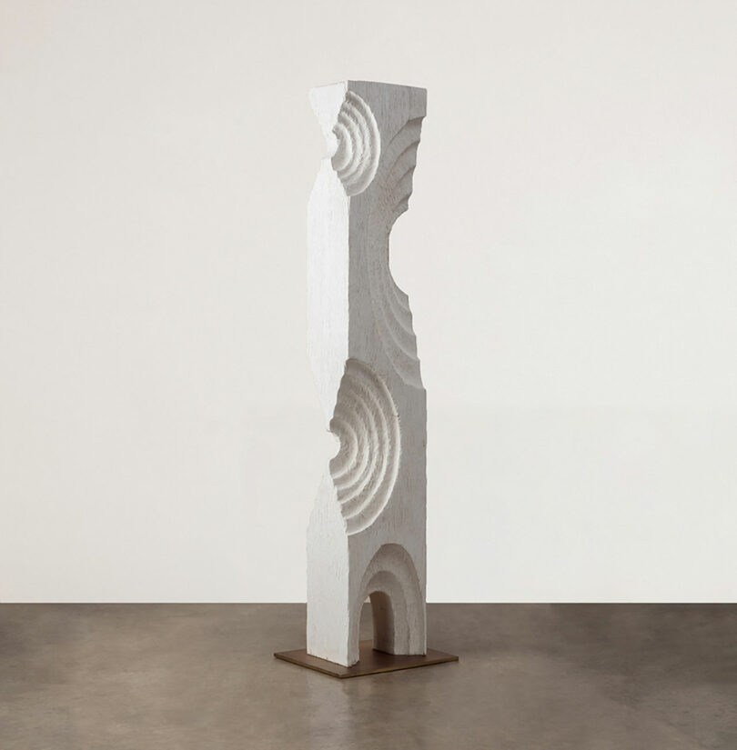 A white sculptural column with concentric circle patterns standing on a metal base in a neutral-toned space.