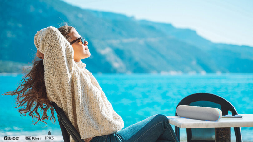 Mujer con gafas de sol y un suéter de punto grande relajándose junto al lago con las montañas al fondo y un altavoz LG StanbyME cerca de una mesa al aire libre