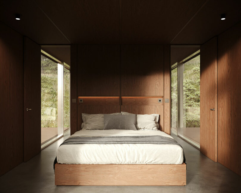 A modern bedroom with a large bed, wooden walls, and floor-to-ceiling windows overlooking a natural landscape.