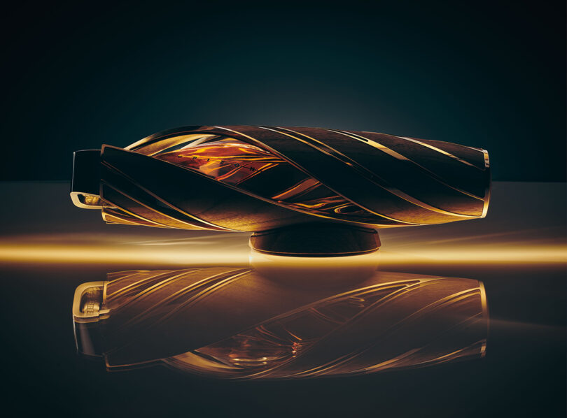 Elegant The Macallan Horizon Whiskey vessel with a spiraling design, backlit to highlight its intricate patterns, displayed on a reflective surface.