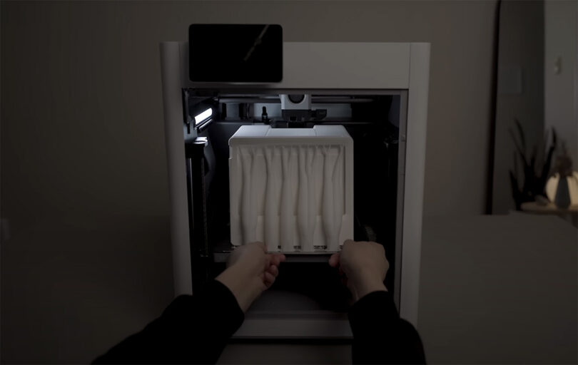 A 3D printer completing a white printed Macintosh Studio dock case from an enclosed printing area using both hands.