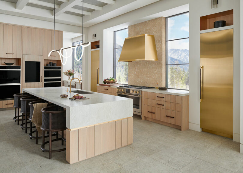 Modern kitchen with wooden cabinets, a central island, brass accents, and a mountain view through large windows, featuring elegant details.