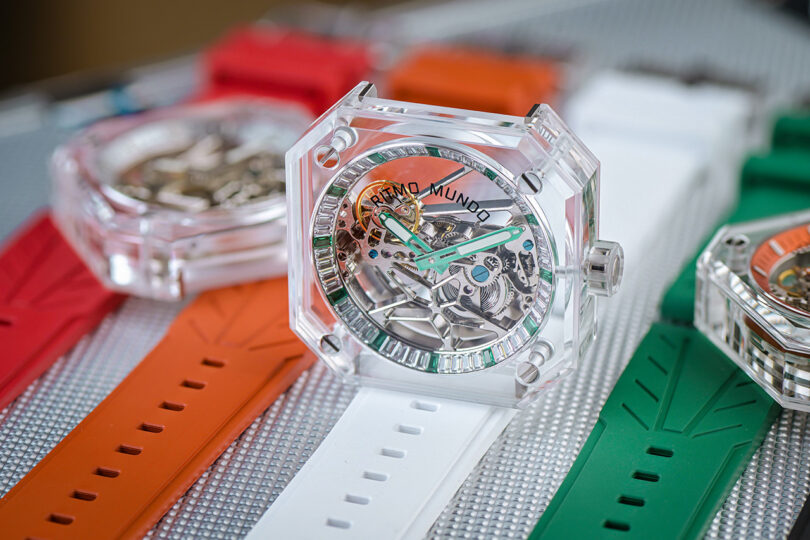 Three Ritmo Mundo Pegasus transparent crystal glass case watches with orange, white, and green silicone wristbands, with watch in the middle set standing on its side vertically.
