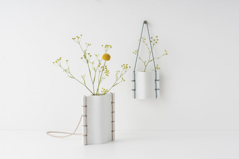 two white ceramic vases with simple foliage hanging on a white wall and on a white table