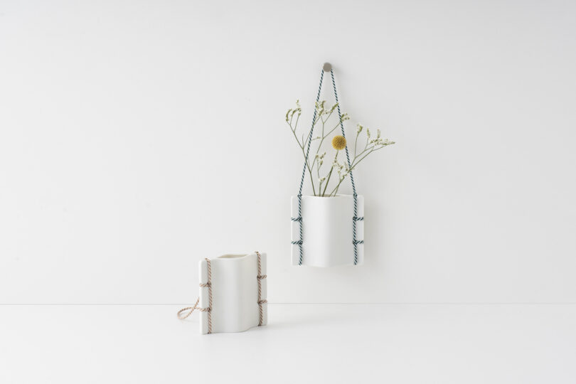 two white ceramic vases with simple foliage hanging on a white wall and on a white table