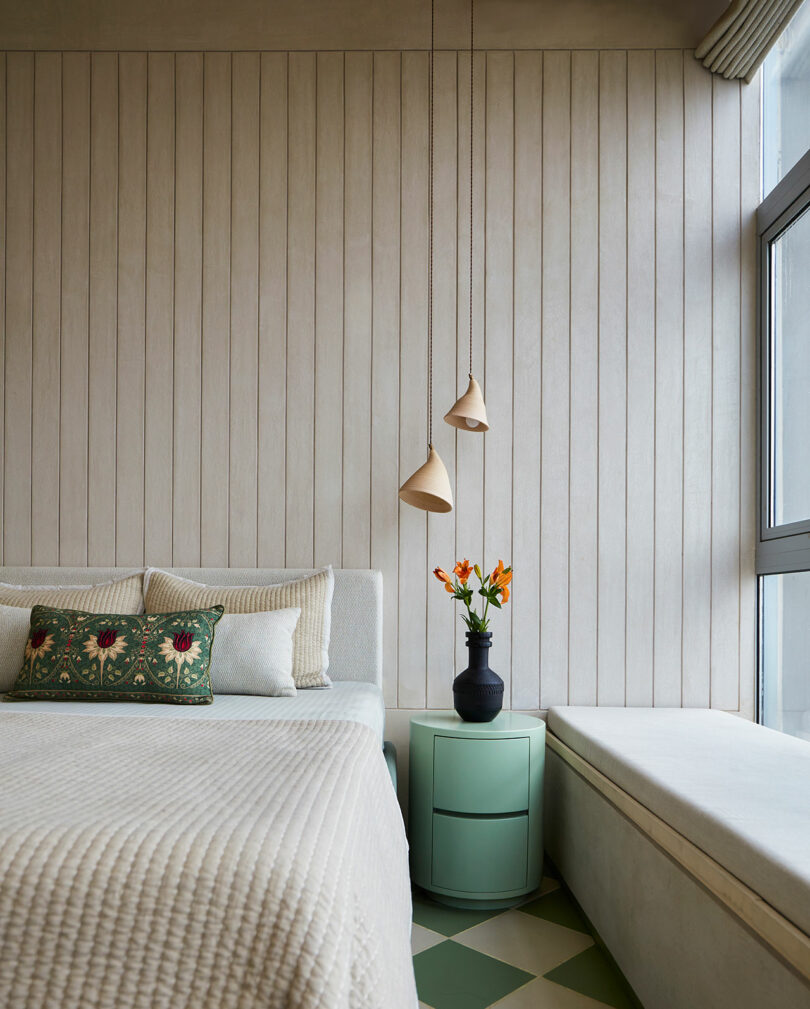 یک اتاق خواب داخلی با یک تخت با بالش های طرح دار، چراغ های آویز، و گلدانی از گل های نارنجی.