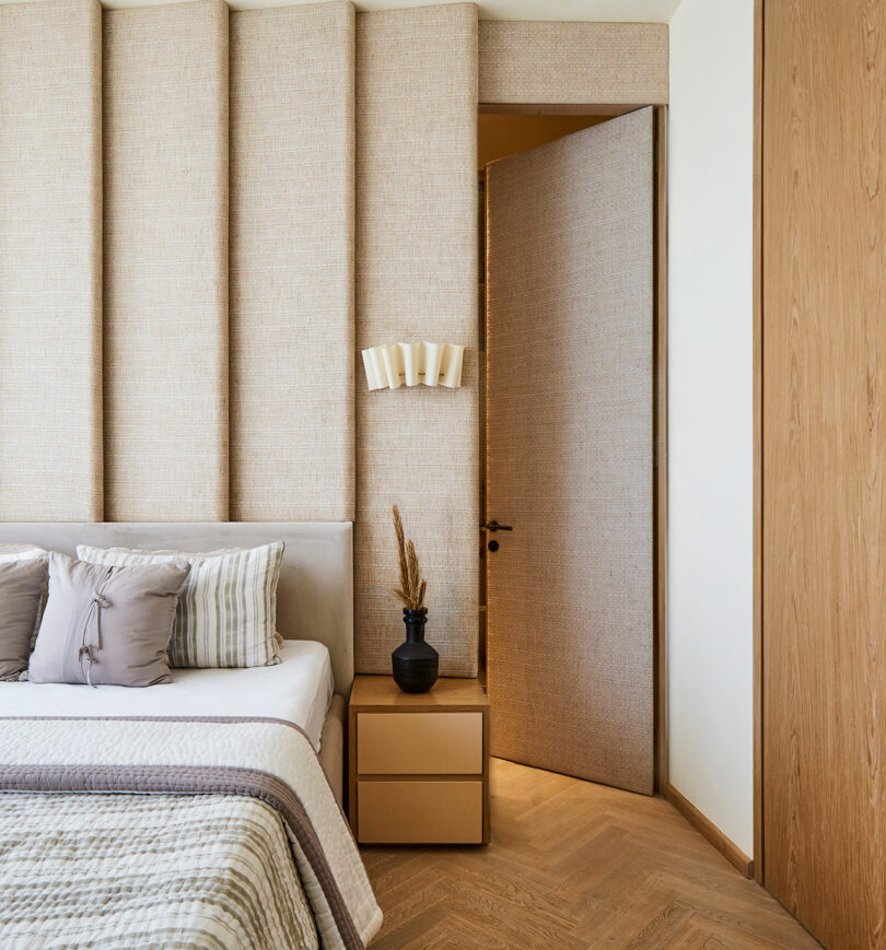یک گوشه اتاق خواب دنج با یک در مخفی که به طور یکپارچه در پوشش دیوار ادغام شده است، در کنار یک تخت خواب منظم با ملافه های بافت دار و یک میز کناری کوچک.