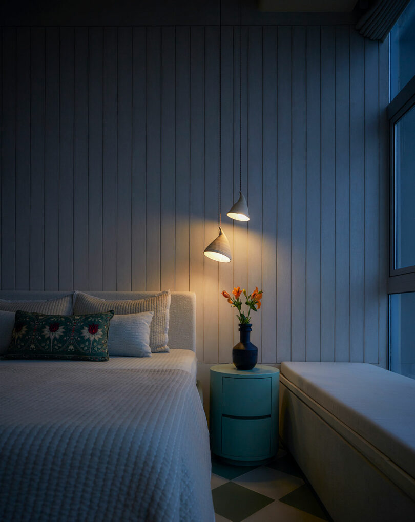 اتاق خوابی آرام در غروب با چراغ آویز روشن و گلدانی از گل های نارنجی روی میز کنار تخت.