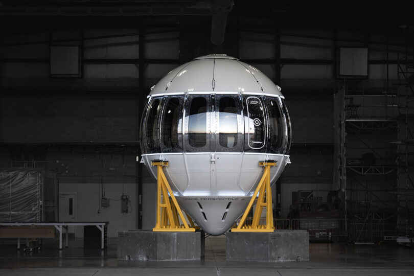 A large white space capsule, part of the world’s first luxury spaceflight service, sitting in a warehouse.