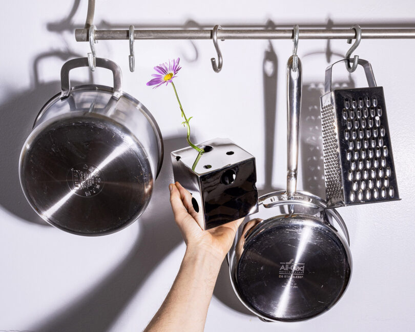 A mirrored cube-shaped vase with a flower in it being held up against a wall of stainless steel pots and pans.