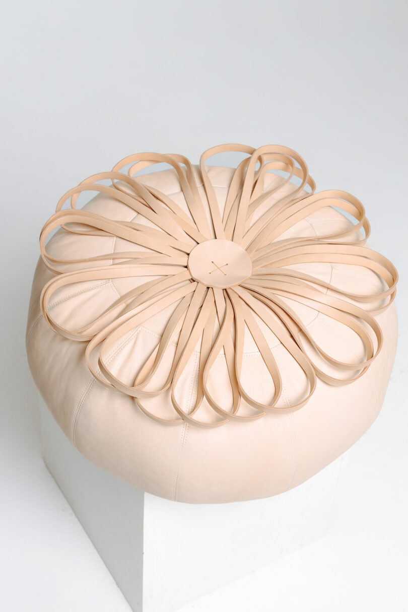Beige ottoman/pouf resembling a blooming flower with petal-like extensions on a white pedestal.
