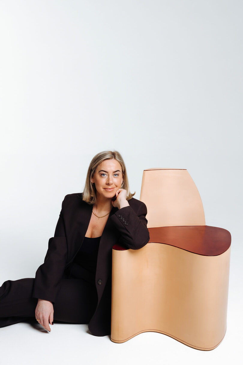 Woman in a black suit sitting next to a large curved wooden structure on a white background.