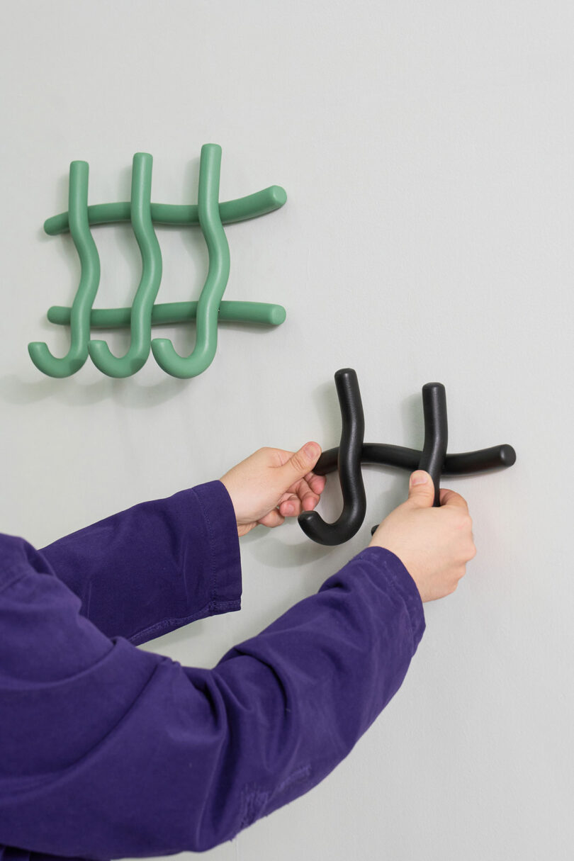 Person hanging a sculptural black hook on a wall next to similar green hooks.