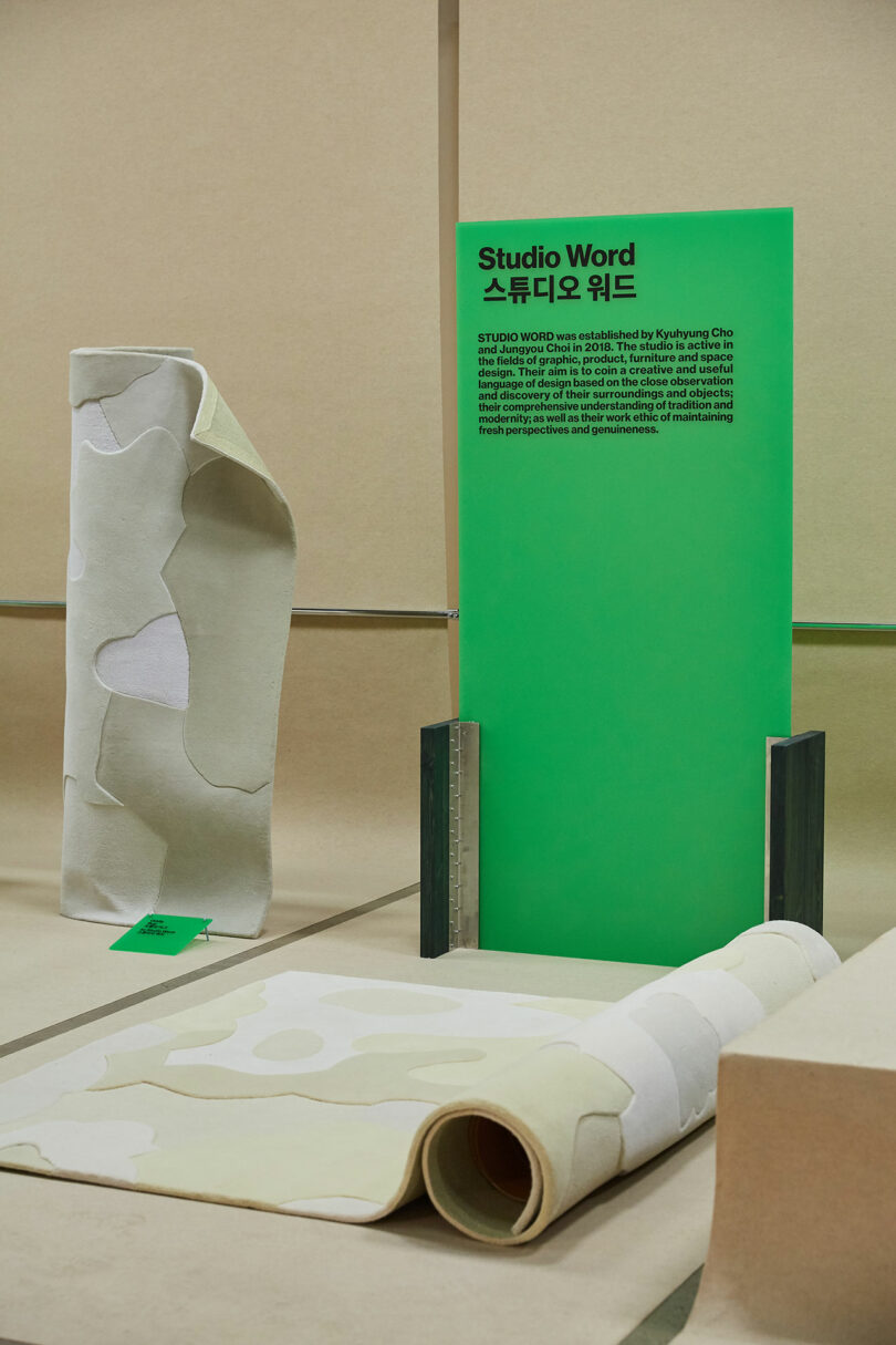 An exhibition space featuring a rolled-up rug with a unique design alongside a standing green informational panel and a sculptural object that echoes the rug's pattern.