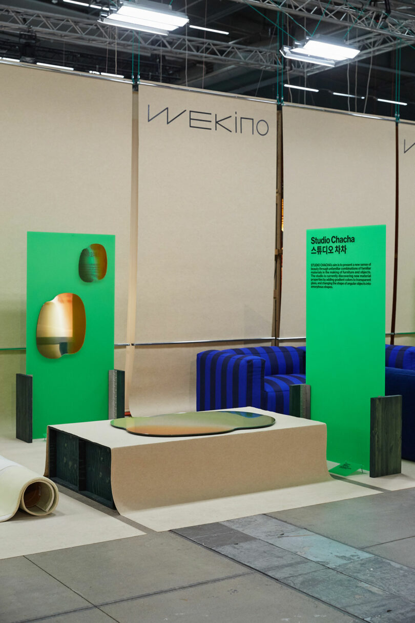 Modern exhibition space with a green backdrop, minimalist furniture, Wekino informational wall text.