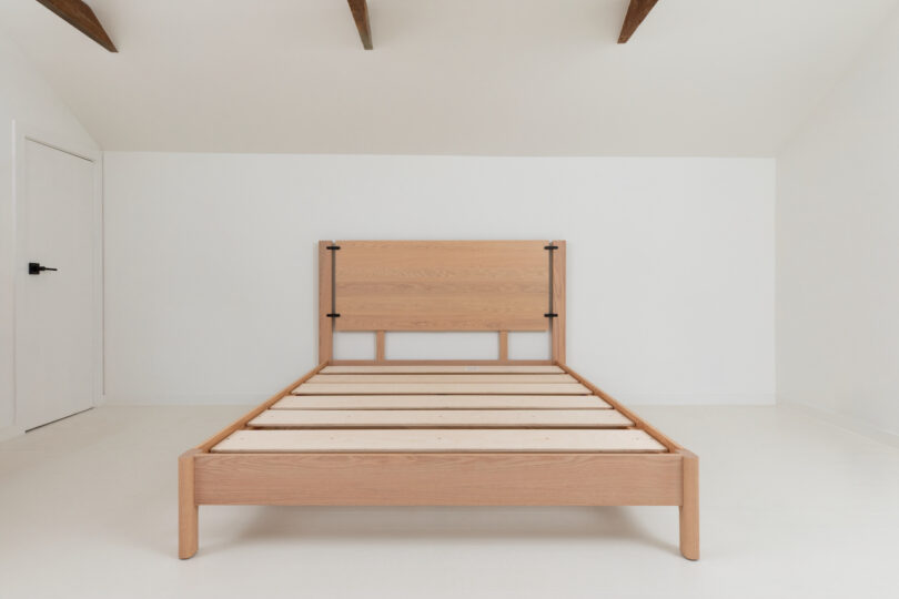 Minimalistic wooden bed frame in a white bedroom with exposed ceiling beams.