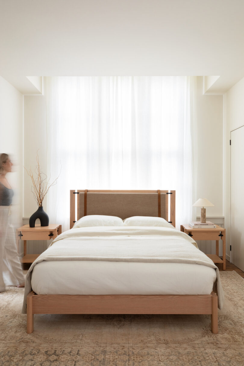 A neatly made bed in a bright room with a person standing by the window.