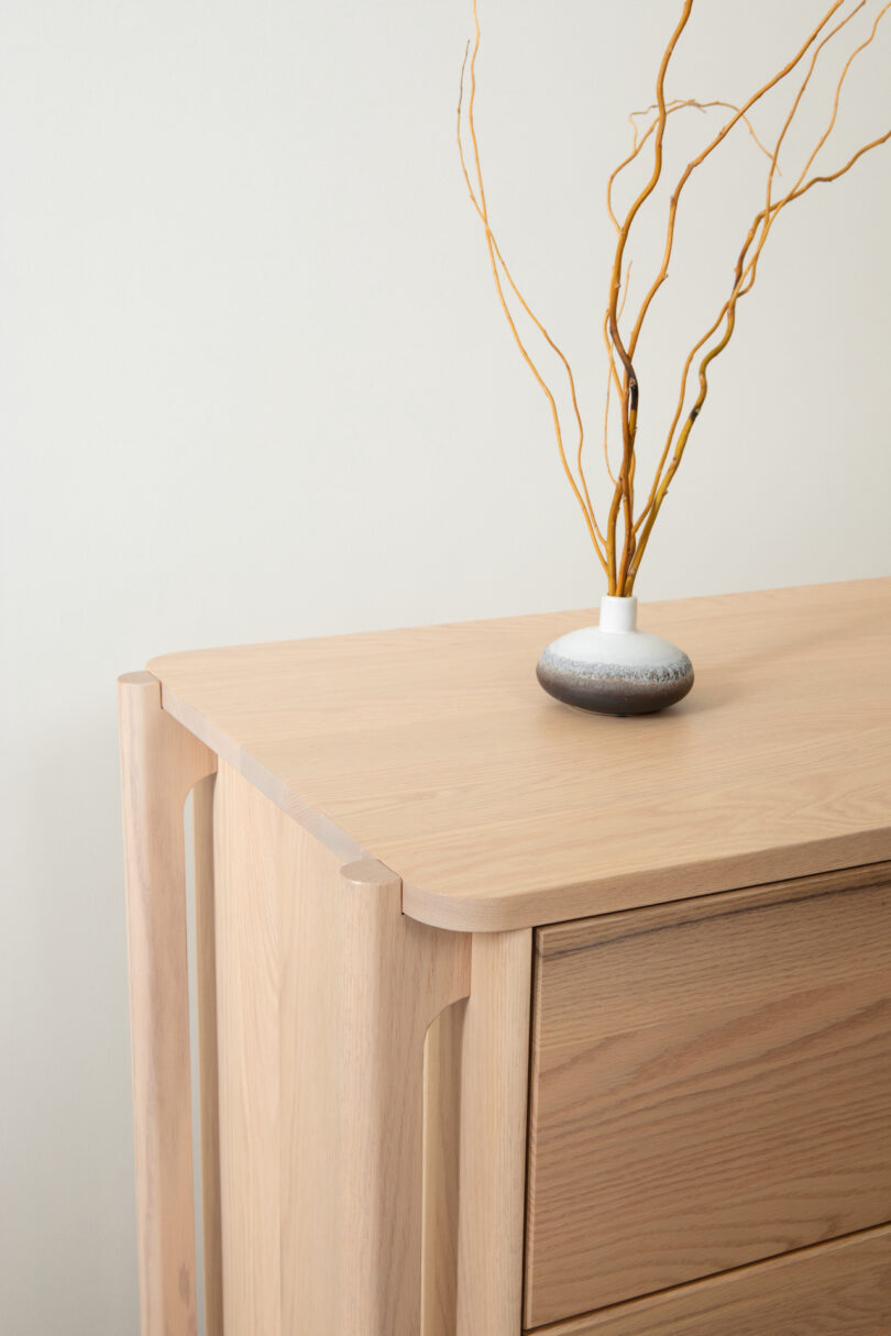 A wooden sideboard with a vase and dried twigs against a neutral background.
