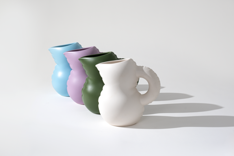 Three colorful ceramic pitchers in blue, green, and white, arranged in a row on a light background with shadows.