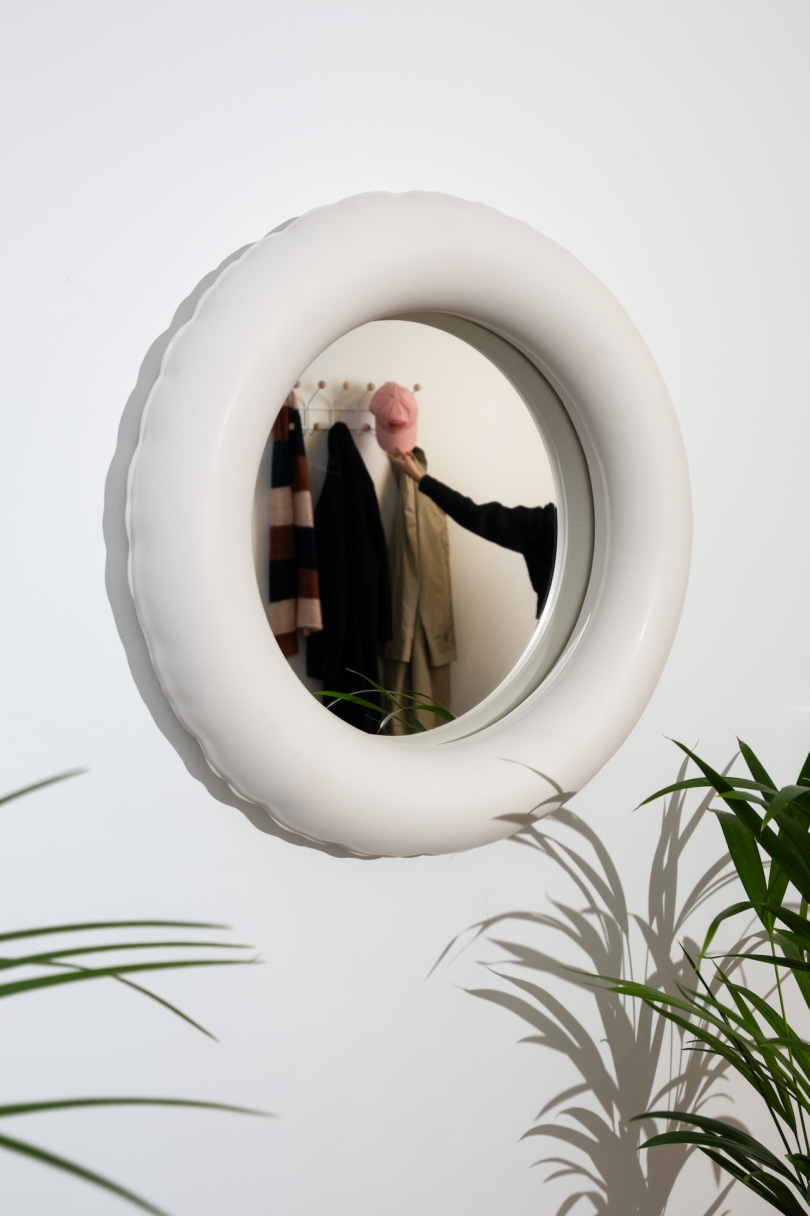 A person's reflection in a round mirror, partially obscured by a potted plant.