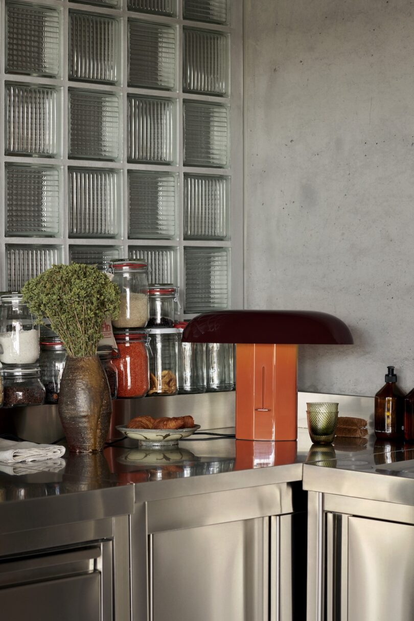 A modern kitchen countertop featuring stainless steel cabinetry, a collection of glass jars with various dry goods, a red toaster, a modern table lamp and a decorative bunch of dried herbs.