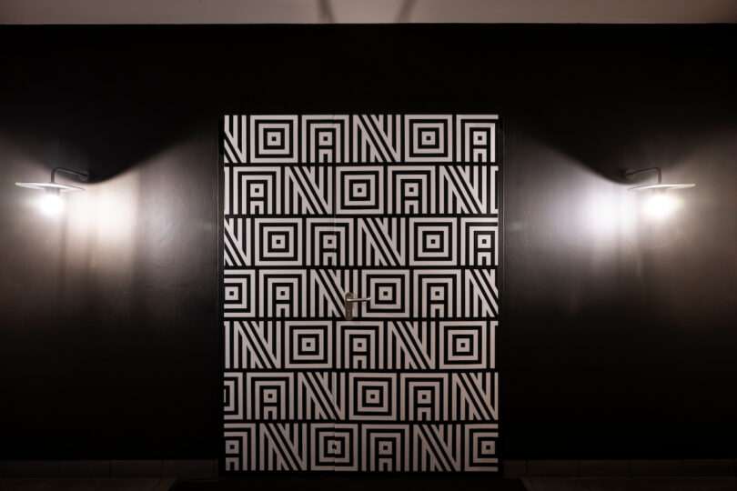 نمای متقارن یک درب NOA با طراحی هنرمندانه با یک الگوی هندسی تکراری، که توسط دو چراغ نصب شده روی دیوار احاطه شده است.
