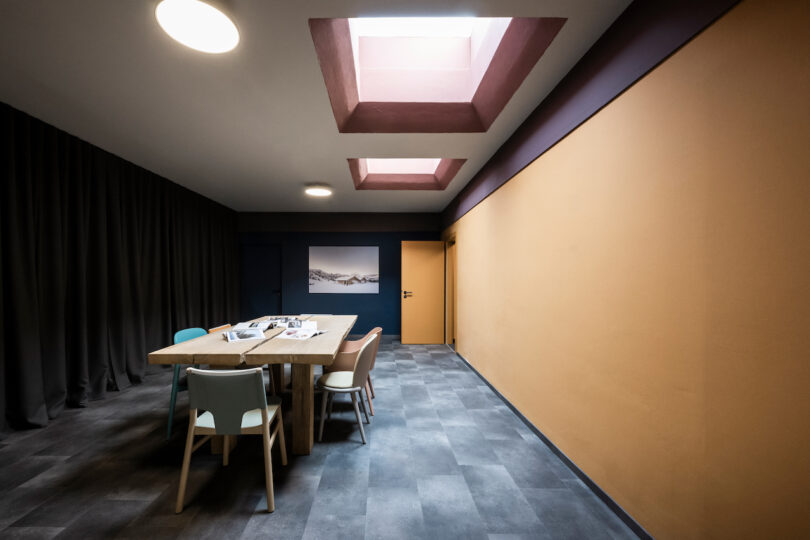 اتاق غذاخوری مدرن با طراحی مینیمالیستی، دارای میز چوبی برای NOA، کف‌پوش خاکستری، یک اثر هنری آبی بزرگ روی دیوار، و نورپردازی بالای سر.