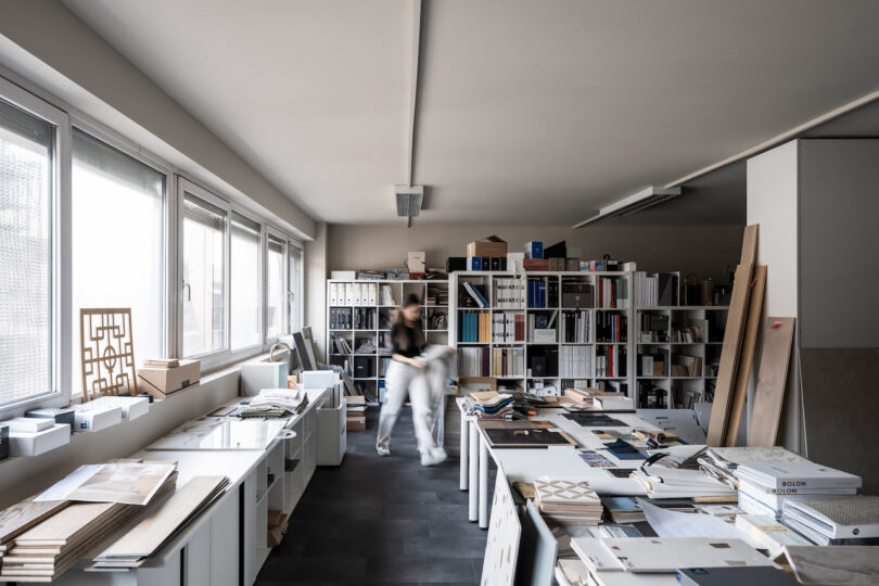 A person is blurred in motion in a busy architectural NOA office filled with bookshelves, desk clutter, and design materials.
