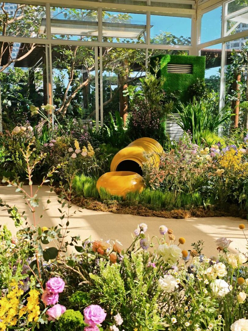 A greenhouse garden featuring yellow structures surrounded by diverse flowers, tall grasses, and dense plant foliage.