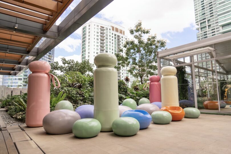 Outdoor terrace with large, colorful abstract sculptures and surrounding greenery against the backdrop of modern high-rise buildings.