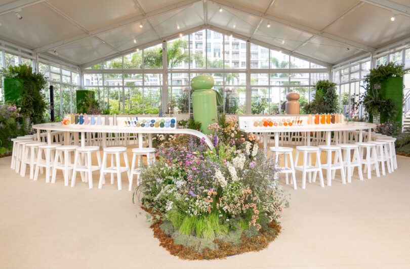 A modern, spacious room with large windows features a long table lined with colorful tableware and surrounded by white stools. The center is adorned with diverse floral arrangements.